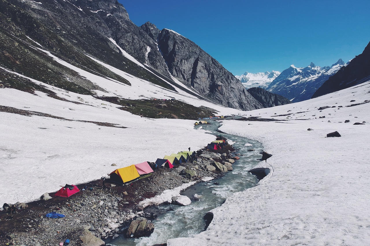 Colorful tents beside the flowing stream of water surrounded by snow covered ground at Balu ka ghera campsite with distant view of Indrasan massif