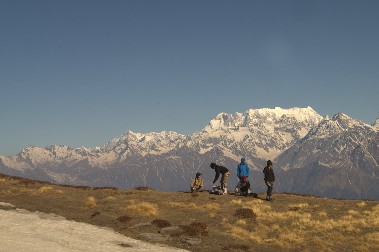 group of 5 people taking rest on the way to kuari pass looking at the snow covered chaukambha massif under the clear blue sky 